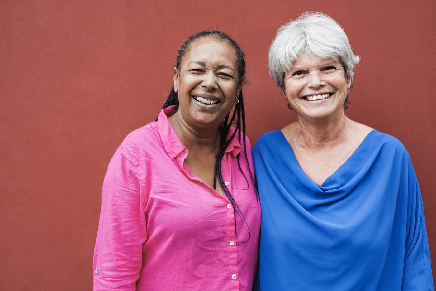 Two older women smiling in front of a red wall, enjoy the benefits of photobiomodulation in Lehi.
